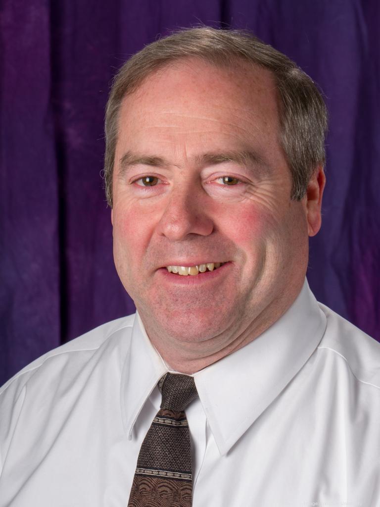A headshot of David Whalen, a middle-aged white man with brown/gray hair, a white shirt and brown patterned tie, and a smile. Purple curtain background.