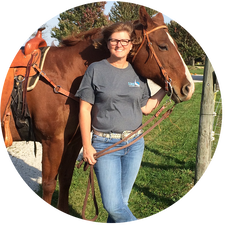 Picture of Tina Felter, a white woman with short brown hair, glasses, and a grey t-shirt. She is standing in front of a large brown horse.