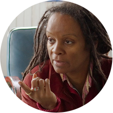 Picture of Veronique Doumbe, an African American woman with dreadlocks looking off to the left and pointing her finger which has a ring on it